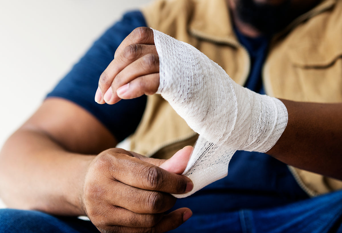 Person with a bandaged hand, wearing a beige jacket and blue jeans, sitting on a white background.