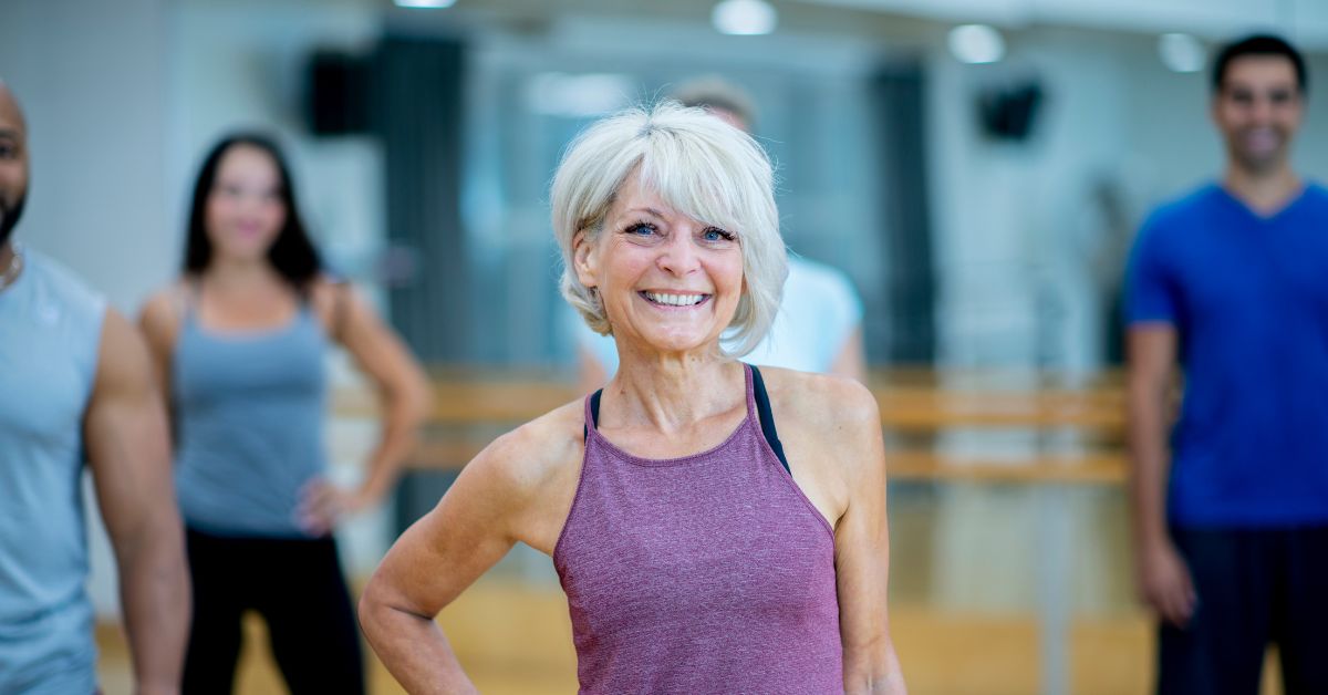 A picture capturing a woman posing for the camera during a group gym class, with other classmates visible in the background.